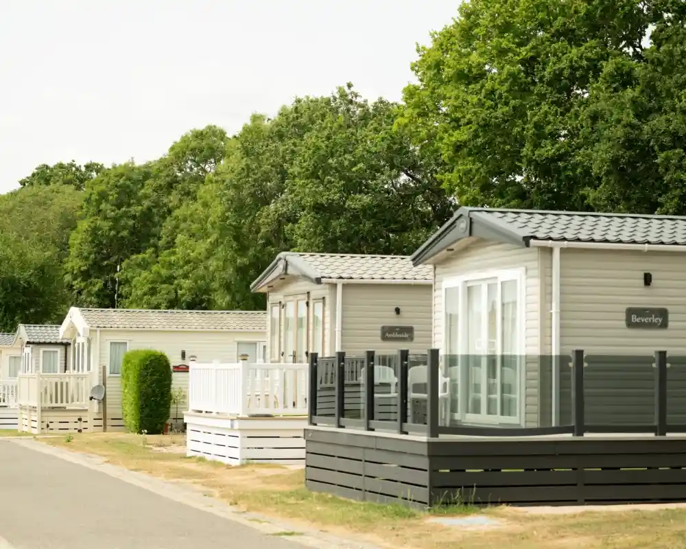 Modern holiday homes with wooden decks line a quiet road, surrounded by lush green trees. The sleek designs feature large windows and varying facades.