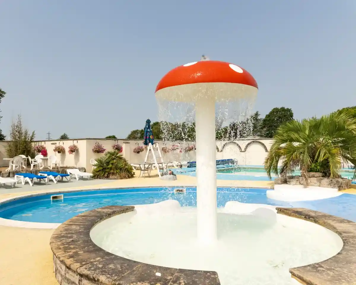 A whimsical fountain shaped like a red and white mushroom, with water cascading down from the top, surrounded by blue swimming pools and lounge chairs. Lush greenery and flower planters are visible in the background under a clear sky.
