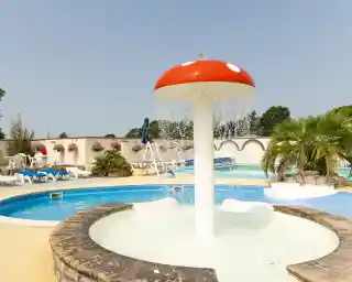 A whimsical fountain shaped like a red and white mushroom, with water cascading down from the top, surrounded by blue swimming pools and lounge chairs. Lush greenery and flower planters are visible in the background under a clear sky.