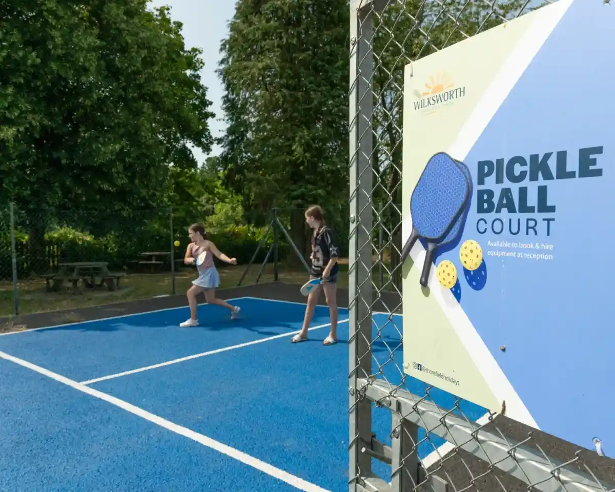 Two young girls playing pickleball on a brightly colored court. A sign nearby indicates that the court is available for booking and mentions equipment rental. Surrounding trees provide a green backdrop.