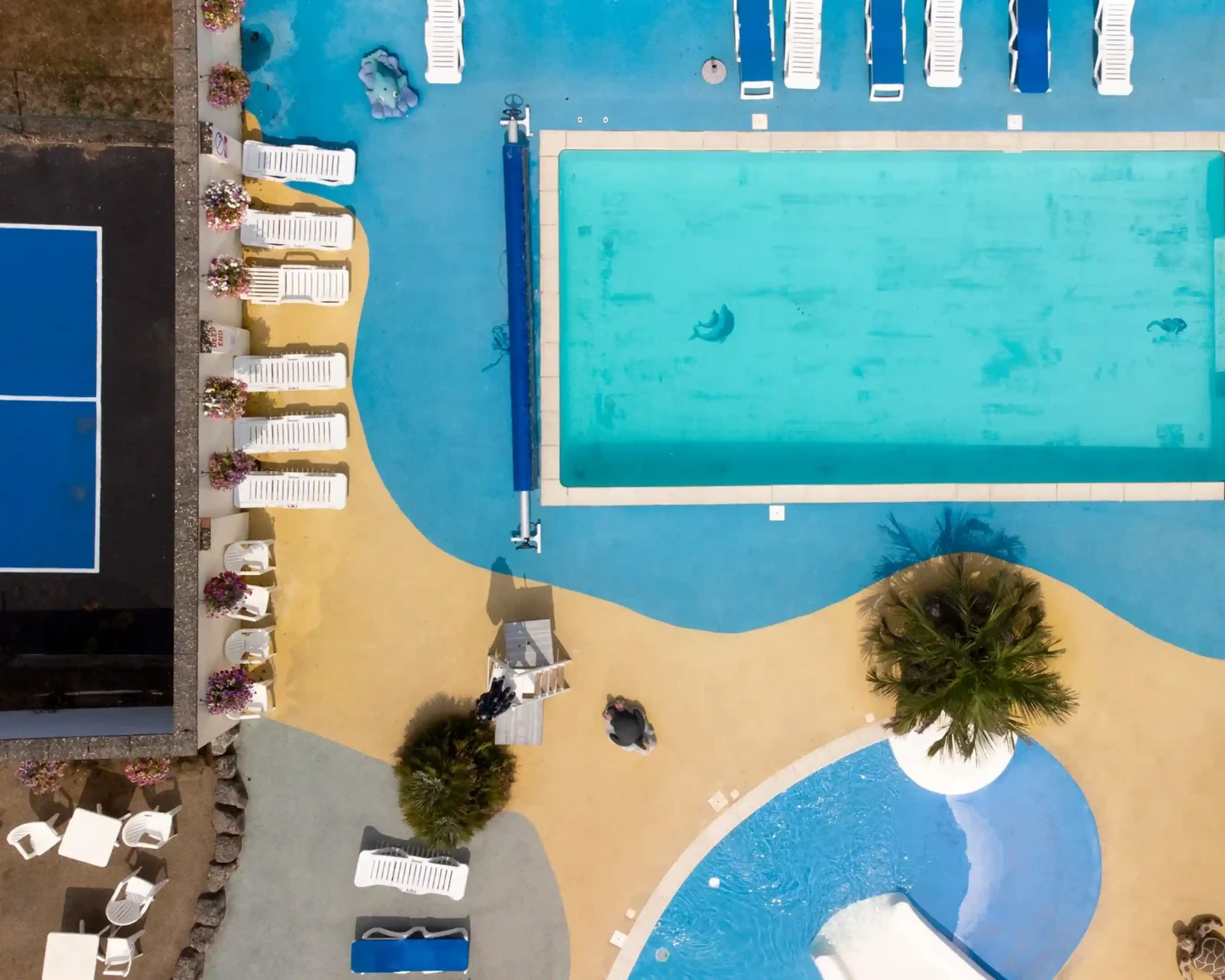 Aerial view of a colorful pool area featuring a large turquoise swimming pool, sun loungers, and a smaller water slide. A tennis court is nearby, with vibrant blue and yellow pathways surrounding the space. Palm trees and flower beds add to the tropical atmosphere.