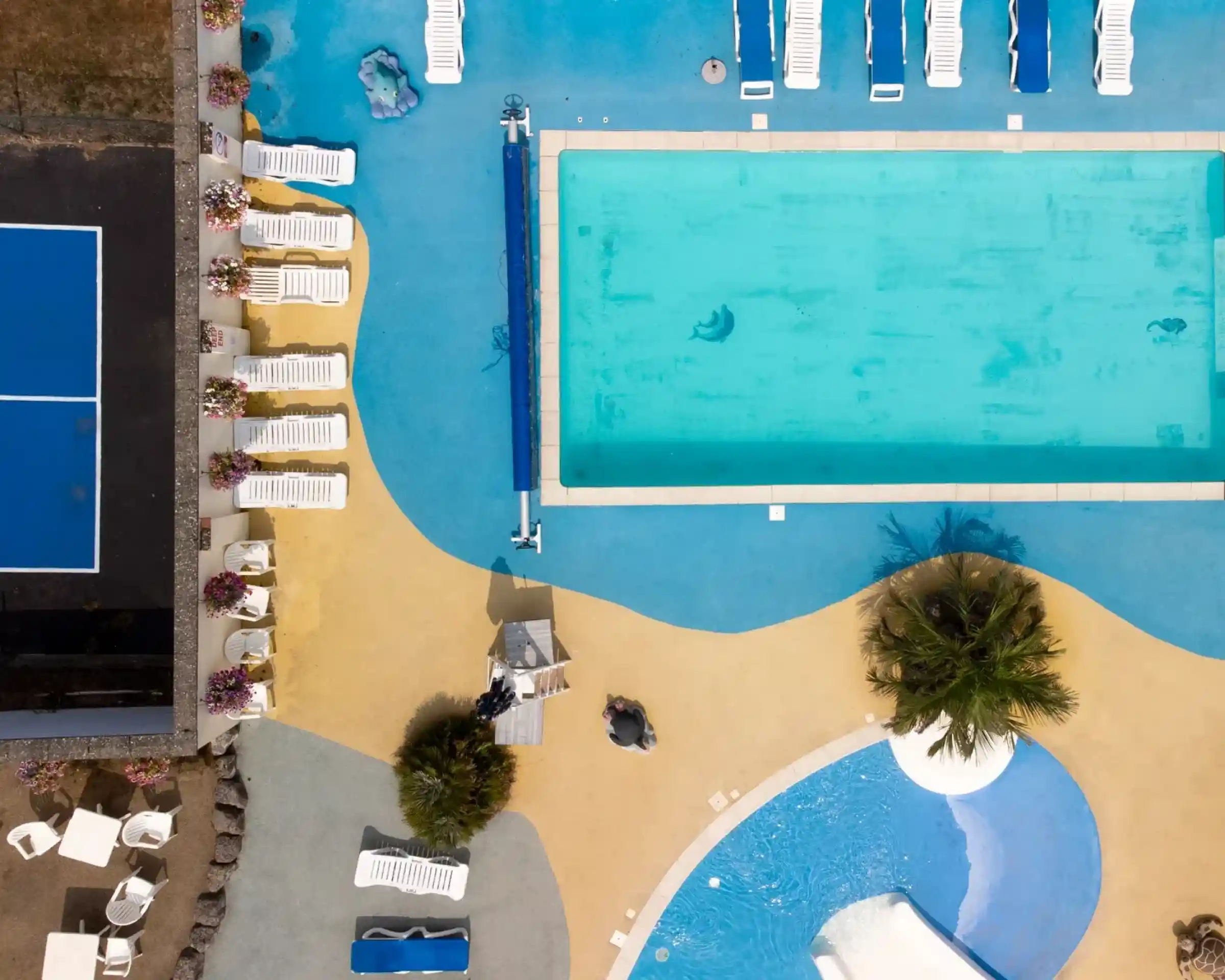 Aerial view of a colorful pool area featuring a large turquoise swimming pool, sun loungers, and a smaller water slide. A tennis court is nearby, with vibrant blue and yellow pathways surrounding the space. Palm trees and flower beds add to the tropical atmosphere.