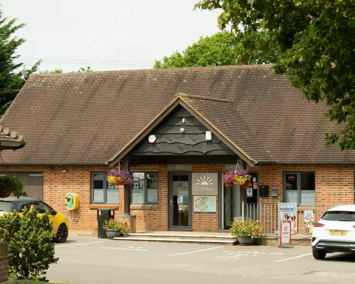A charming brick building with a sloped roof, featuring large windows and hanging flower baskets. The entrance is framed by plants, and there are signs indicating services and parking. A white car is parked nearby.