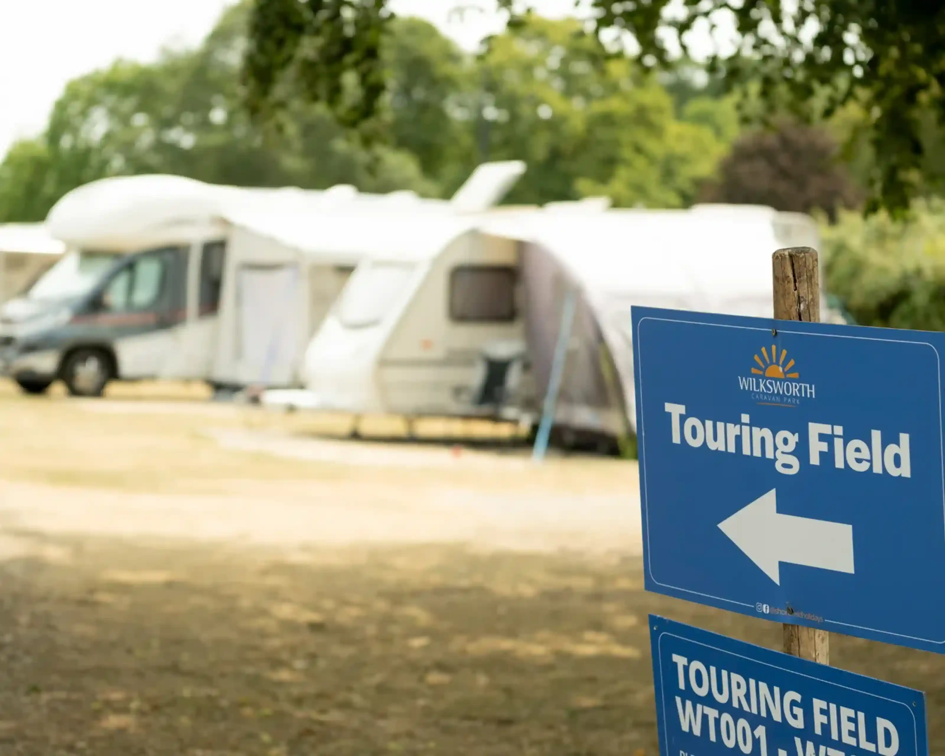 A blue sign pointing left indicates the touring field at a campsite, with several caravans and vehicles set up in the background amidst trees and grass.