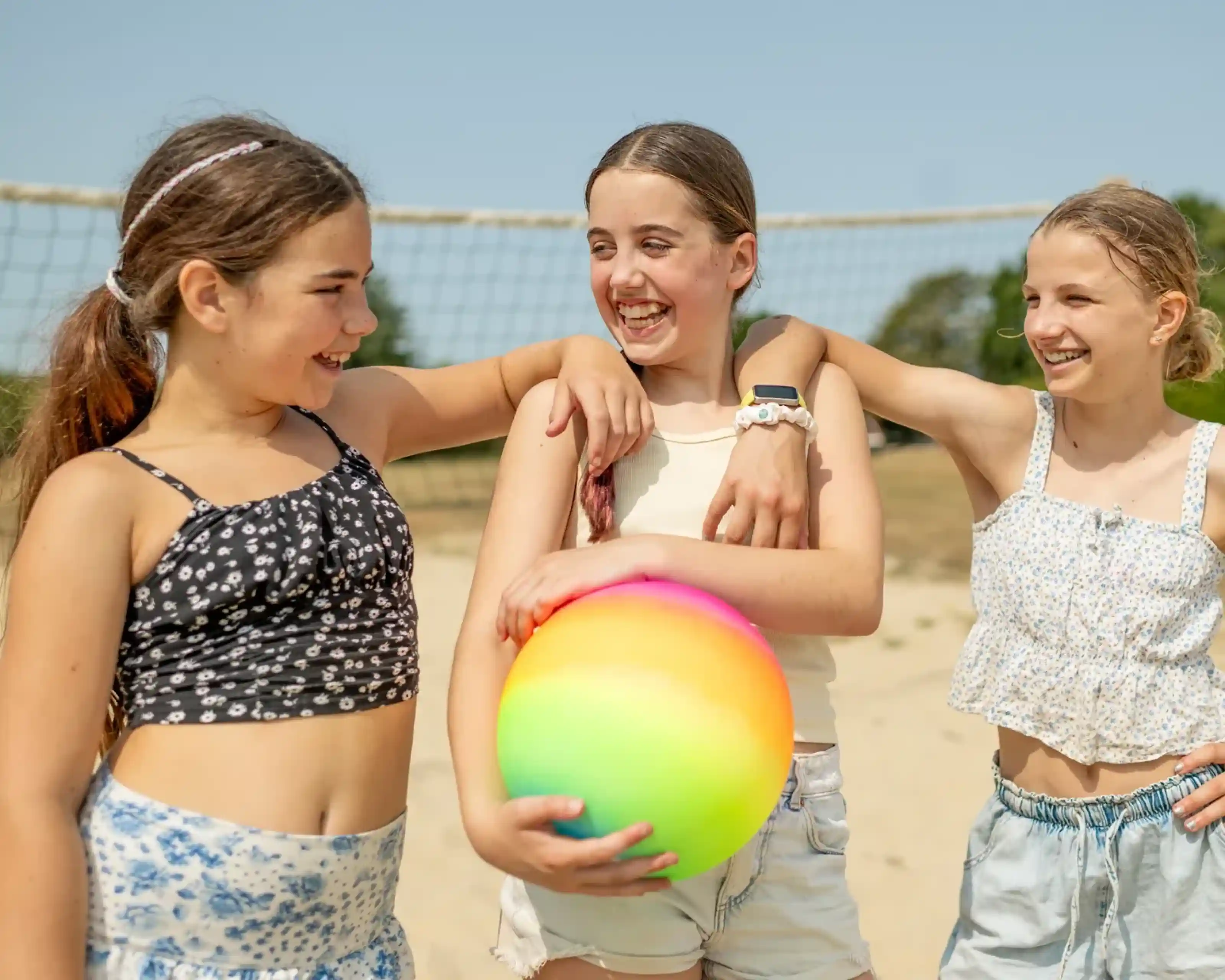 Three girls are smiling and laughing together on a sandy beach, holding a colorful beach ball. They are dressed in light summer outfits, with a volleyball net visible in the background. The sun is shining brightly.