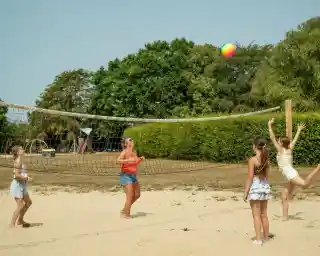 Four children play beach volleyball on a sandy court. One child jumps to hit a colorful beach ball while others watch. Lush greenery and playground equipment are visible in the background.