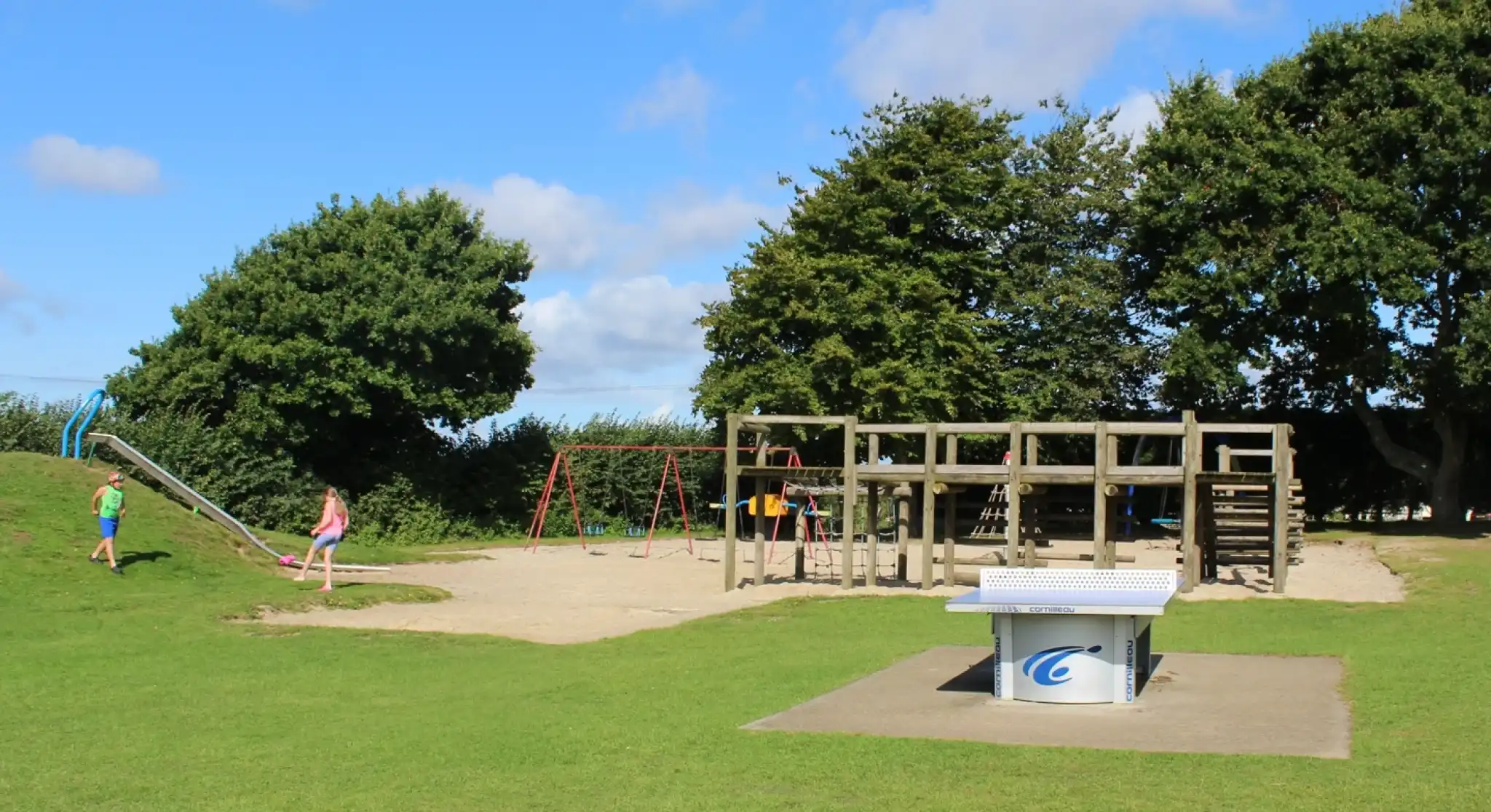 A children's playground featuring a wooden play structure, swings, and a slide, surrounded by grassy areas and trees under a blue sky with fluffy clouds.