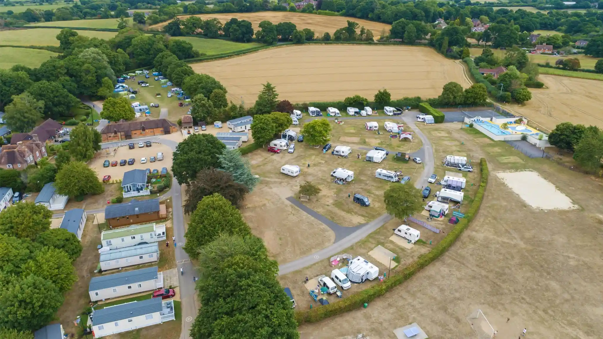 An aerial view of a campsite surrounded by lush green fields. The campsite features several caravans, mobile homes, and tents arranged along winding paths. Trees provide shade throughout the area, while vehicles are parked nearby. The landscape includes golden fields and patches of forest in the background.