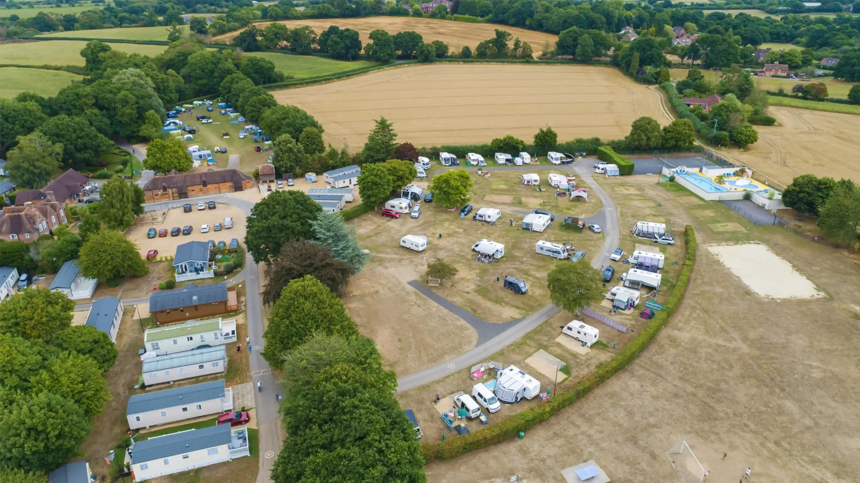 An aerial view of a campsite surrounded by lush green fields. The campsite features several caravans, mobile homes, and tents arranged along winding paths. Trees provide shade throughout the area, while vehicles are parked nearby. The landscape includes golden fields and patches of forest in the background.