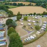 An aerial view of a campsite surrounded by lush green fields. The campsite features several caravans, mobile homes, and tents arranged along winding paths. Trees provide shade throughout the area, while vehicles are parked nearby. The landscape includes golden fields and patches of forest in the background.