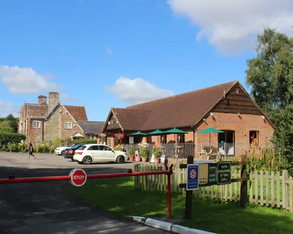 A brick building with a sloped roof sits in a rural area, surrounded by greenery. Several cars are parked in front, and outdoor seating is available under umbrellas. A stop sign is visible at the entrance, along with informational signs. Blue sky with clouds overhead.