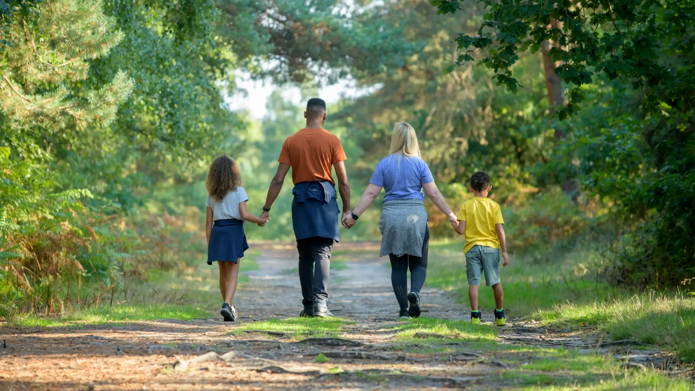 A family of four walks hand in hand along a dirt path in a lush, green forest. Two children, a girl with curly hair and a boy wearing a yellow shirt, are accompanied by an adult male and an adult female. They are all facing away from the camera and enjoying a sunny day outdoors.