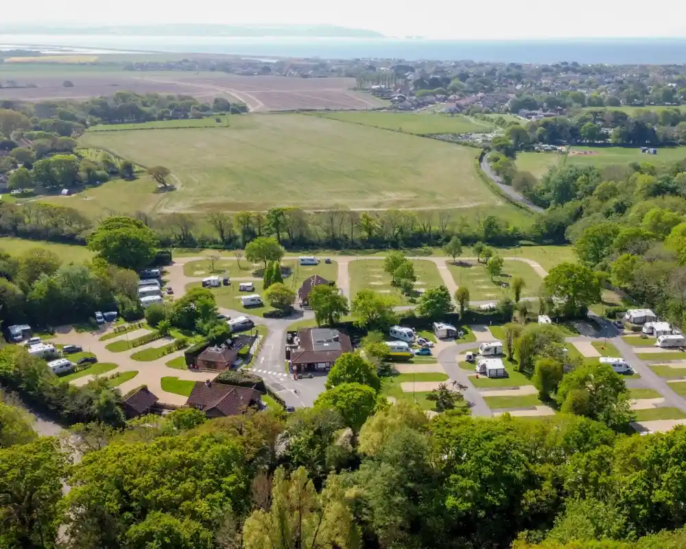 Aerial view of a camping area surrounded by lush greenery. The site features several caravans and tents arranged in neat rows. In the background, fields and a distant view of a coastline can be seen.