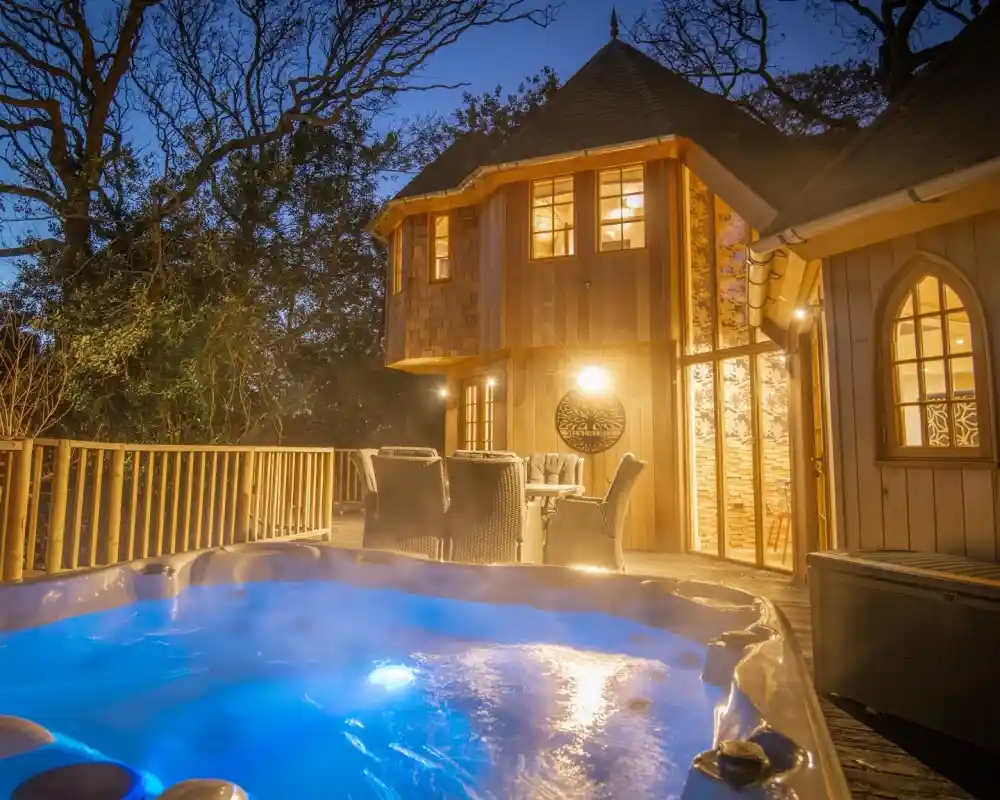 A cozy wooden cabin illuminated at dusk, featuring large windows and a deck with seating. In the foreground, a hot tub emits blue light, surrounded by trees.