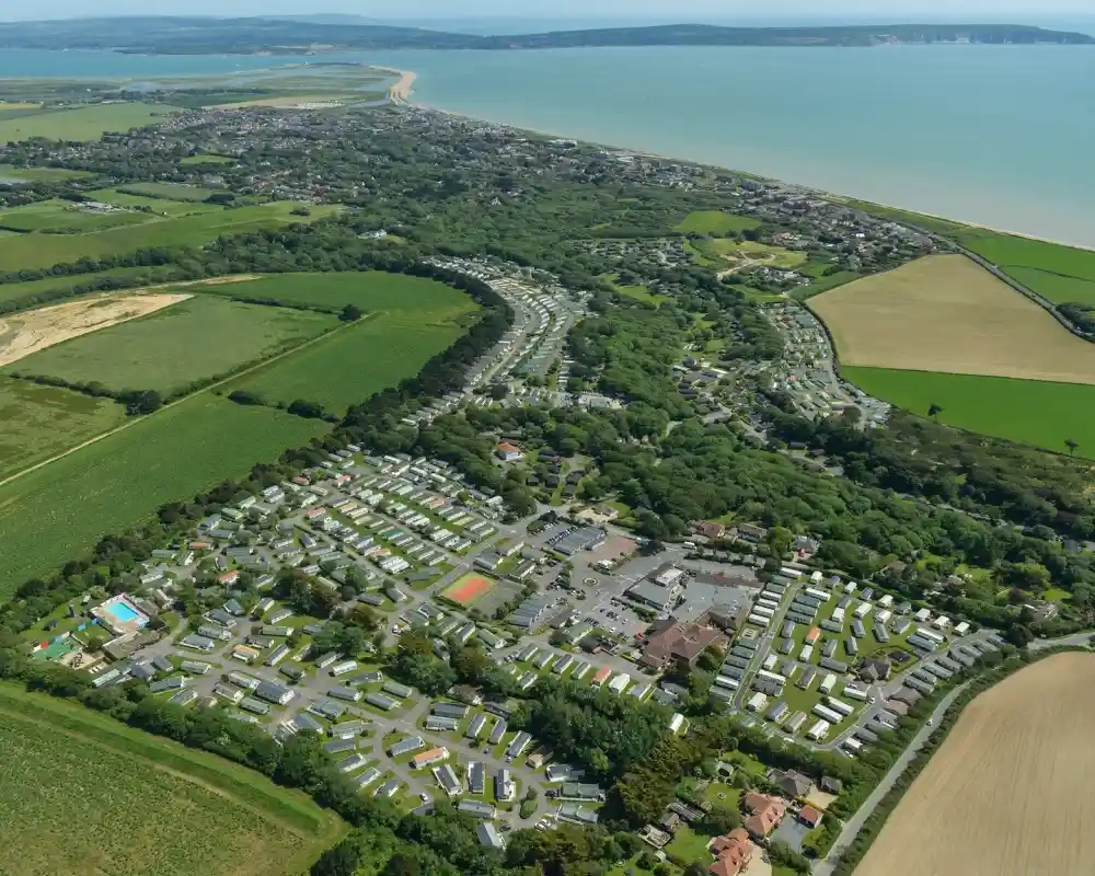 Aerial view of a coastal holiday park surrounded by green fields, showcasing rows of caravan accommodations and amenities. The coastline stretches in the background, with a neighboring town visible along the shore.