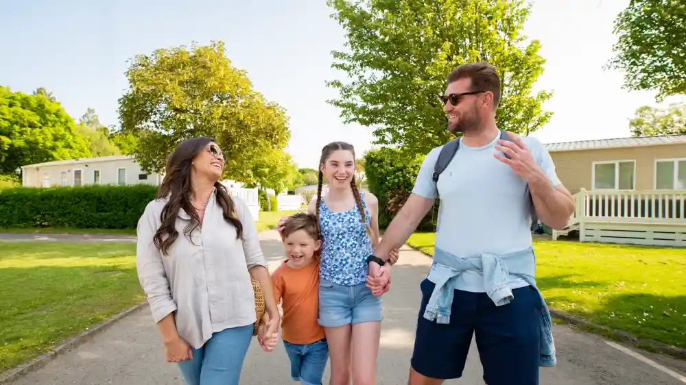 A family of four enjoys a sunny day outdoors, walking along a pathway. The mother and father are smiling and holding hands with their two children, a boy and a girl. The boy, in an orange shirt, playfully runs ahead, while the girl, wearing a blue top, walks beside the mother. Lush greenery and holiday homes are visible in the background.