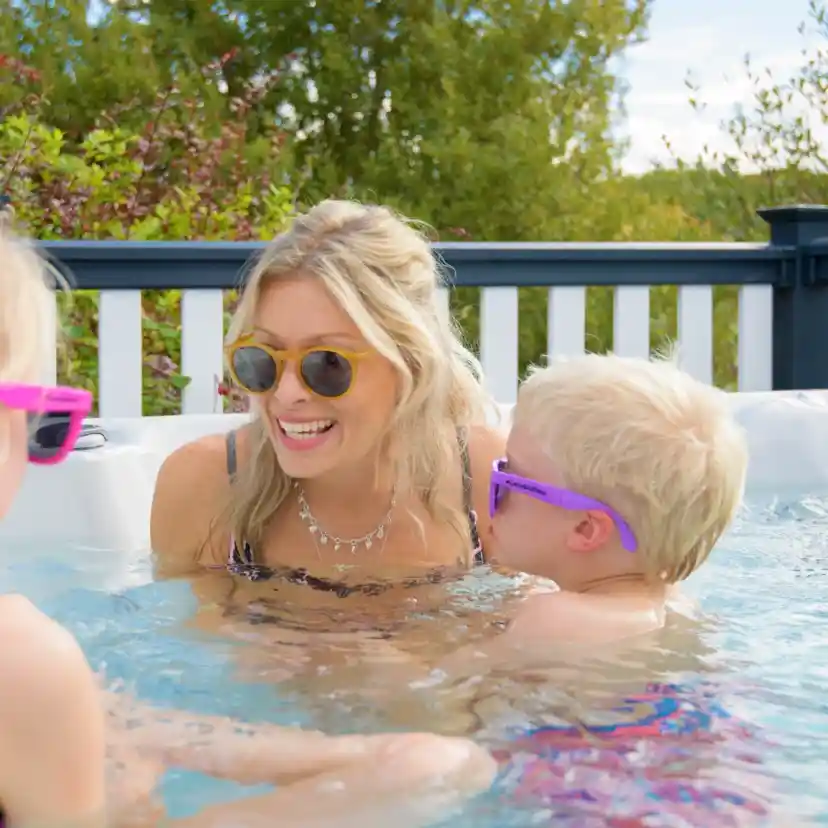 A woman in sunglasses enjoys a lively moment in a hot tub with two children, also wearing sunglasses. They are smiling and splashing water, surrounded by greenery and a blue sky.