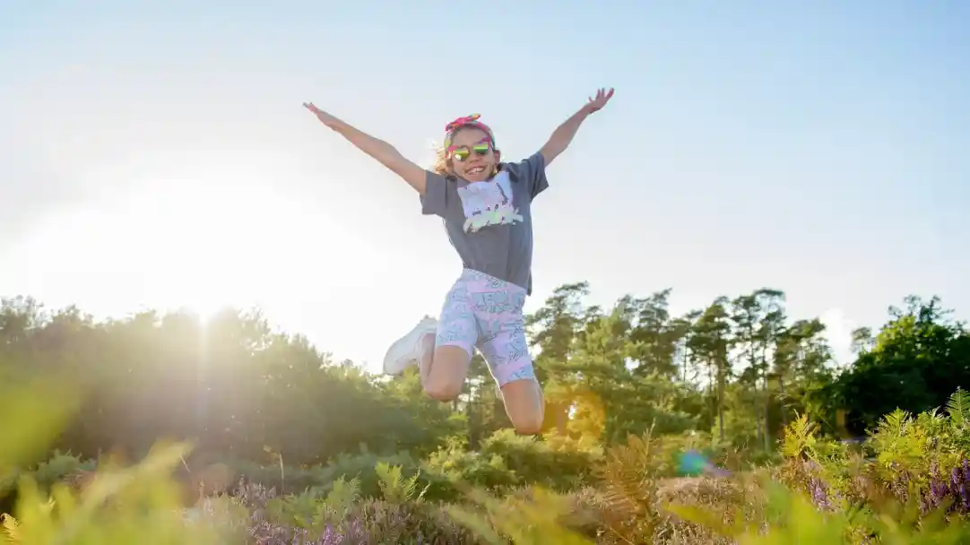 A young girl jumps joyfully in a sunny outdoor setting, wearing a grey t-shirt, colourful shorts, and sunglasses. The background features trees and colourful plants, with sunlight creating a bright, cheerful atmosphere.