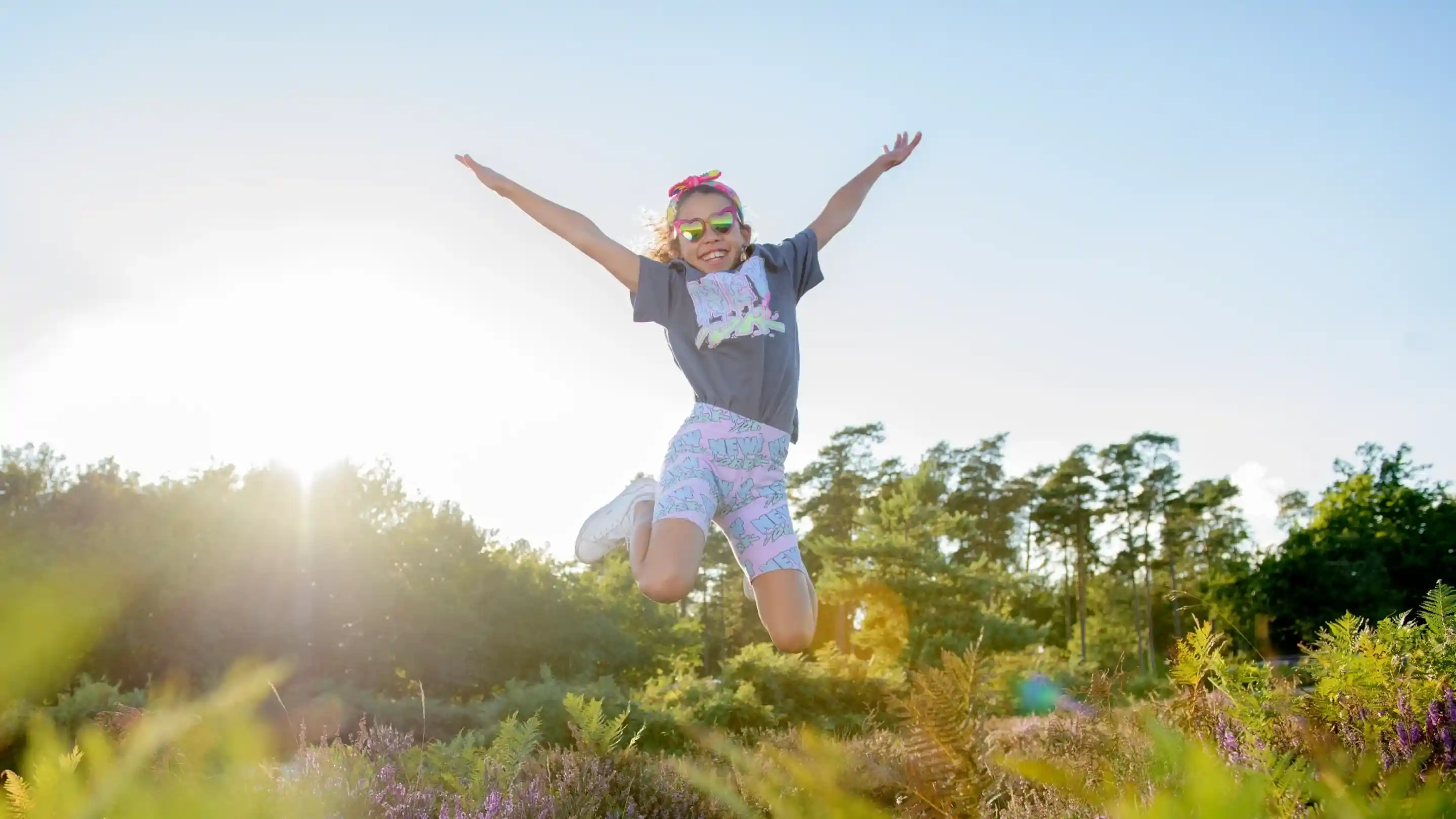 A young girl jumps joyfully in a sunny outdoor setting, wearing a gray t-shirt, colorful shorts, and sunglasses. The background features trees and colorful plants, with sunlight creating a bright, cheerful atmosphere.