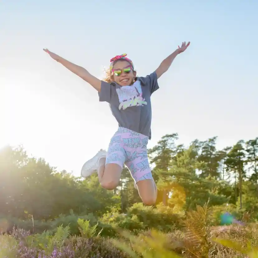 A young girl jumps joyfully in a sunny outdoor setting, wearing a grey t-shirt, colourful shorts, and sunglasses. The background features trees and colourful plants, with sunlight creating a bright, cheerful atmosphere.