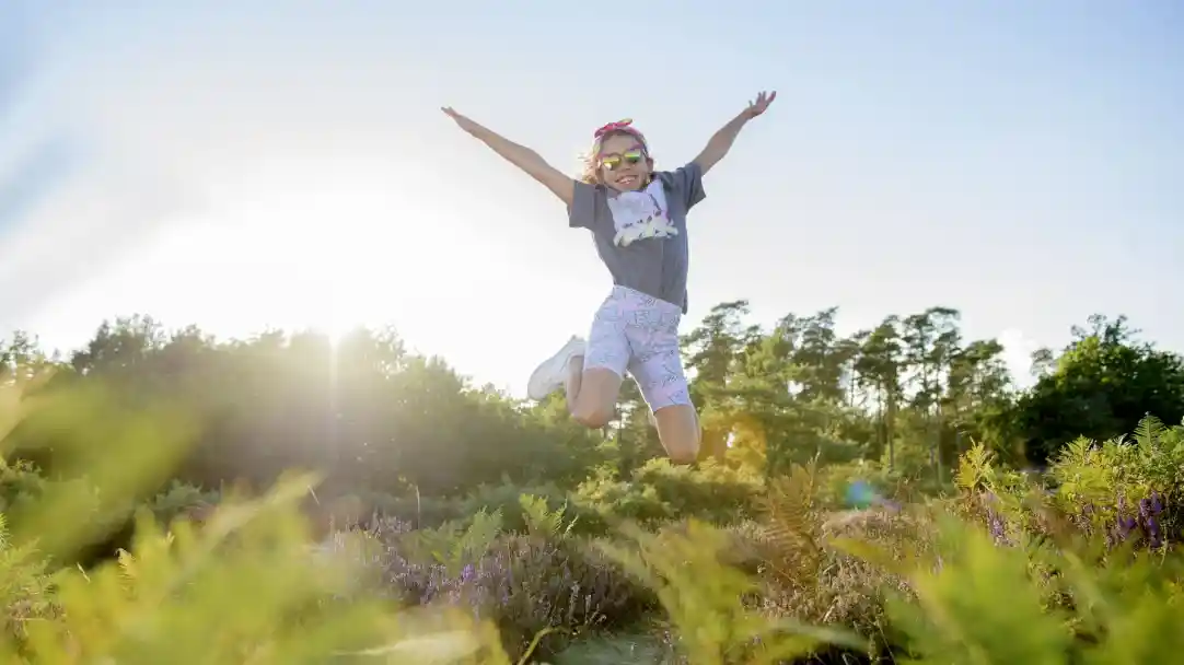 A young girl jumps joyfully in a sunlit field, wearing sunglasses and a colorful outfit. Green foliage and flowers surround her, creating a vibrant and cheerful atmosphere.