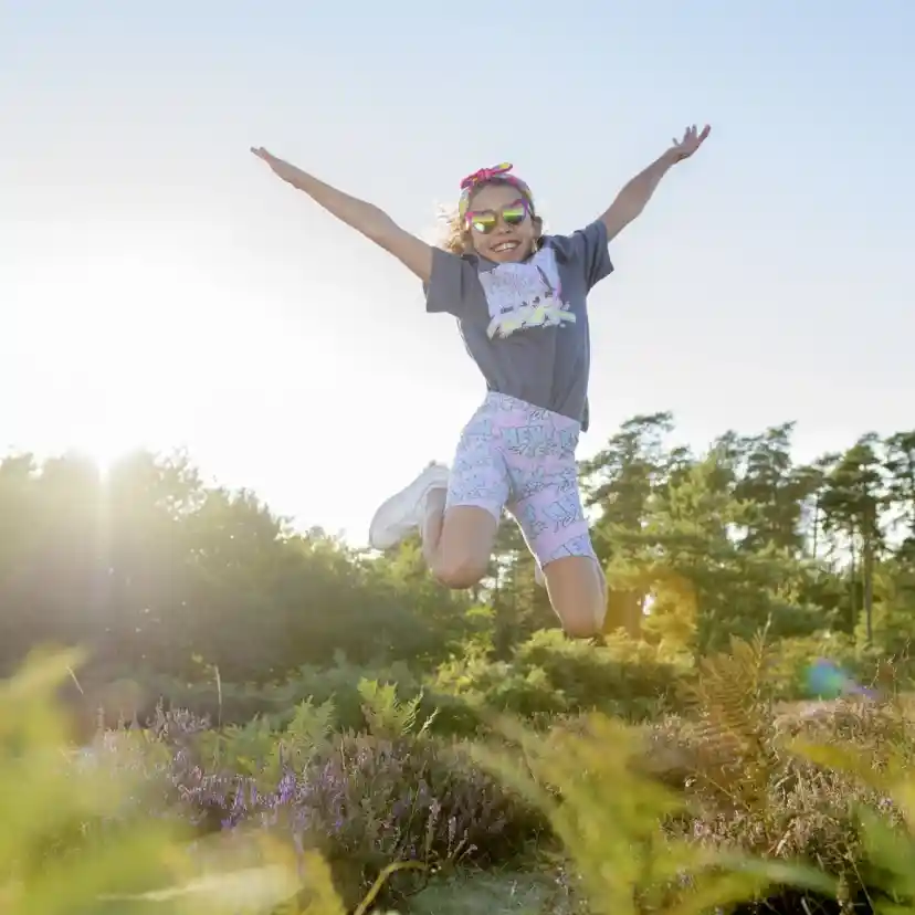 A young girl jumps joyfully in a sunlit field, wearing sunglasses and a colorful outfit. Green foliage and flowers surround her, creating a vibrant and cheerful atmosphere.