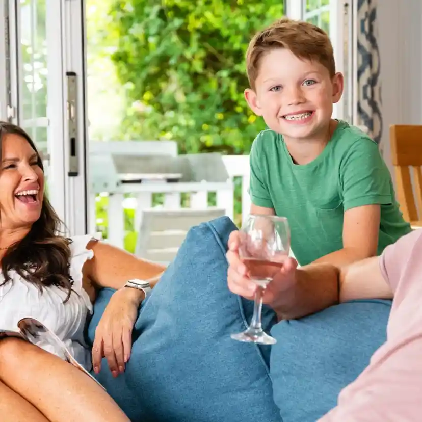 A woman and a man relax on a couch, smiling and laughing, while a young boy stands between them with a big grin. The setting is a bright, cozy living room with greenery visible outside. The woman is holding a magazine, and the man has a glass in his hand.