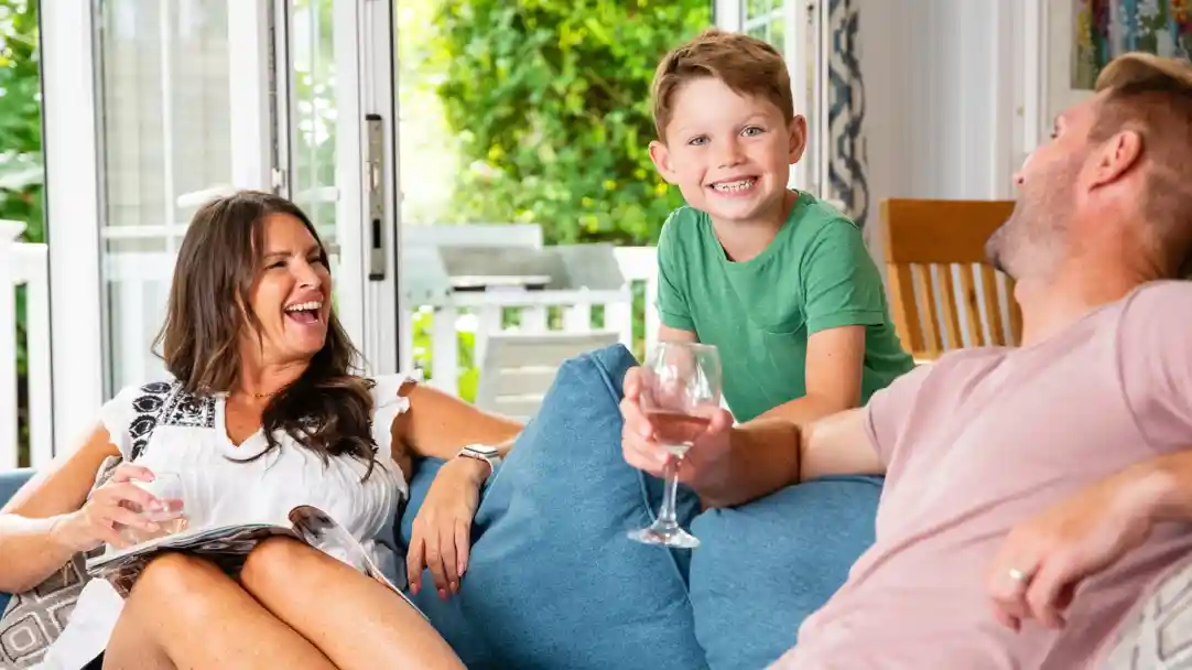 A woman and a man relax on a couch, smiling and laughing, while a young boy stands between them with a big grin. The setting is a bright, cozy living room with greenery visible outside. The woman is holding a magazine, and the man has a glass in his hand.