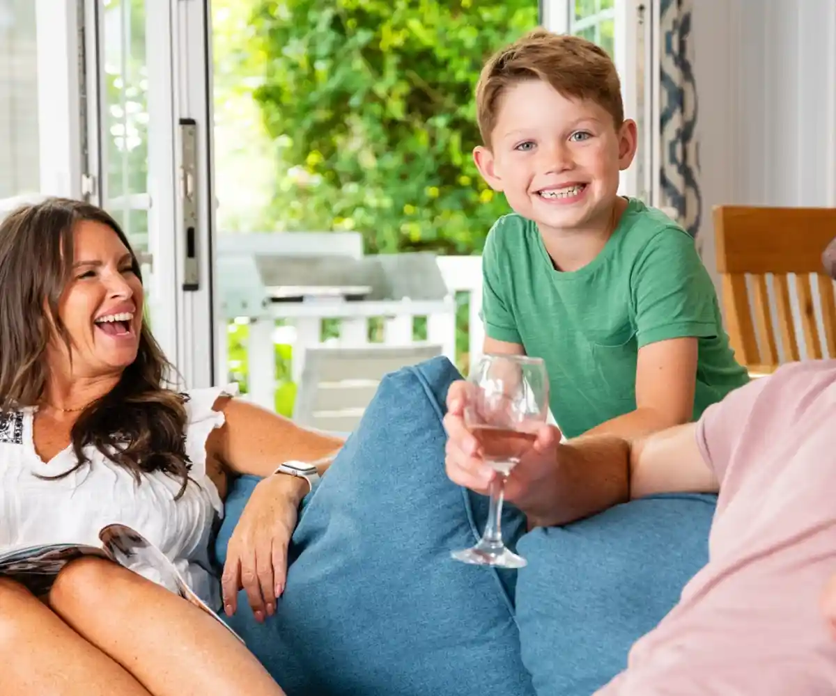 A couple stands on a balcony with a modern home in the background. The man and woman are smiling, dressed casually, with green grass and a landscaped area surrounding the house. The sky is clear and blue.