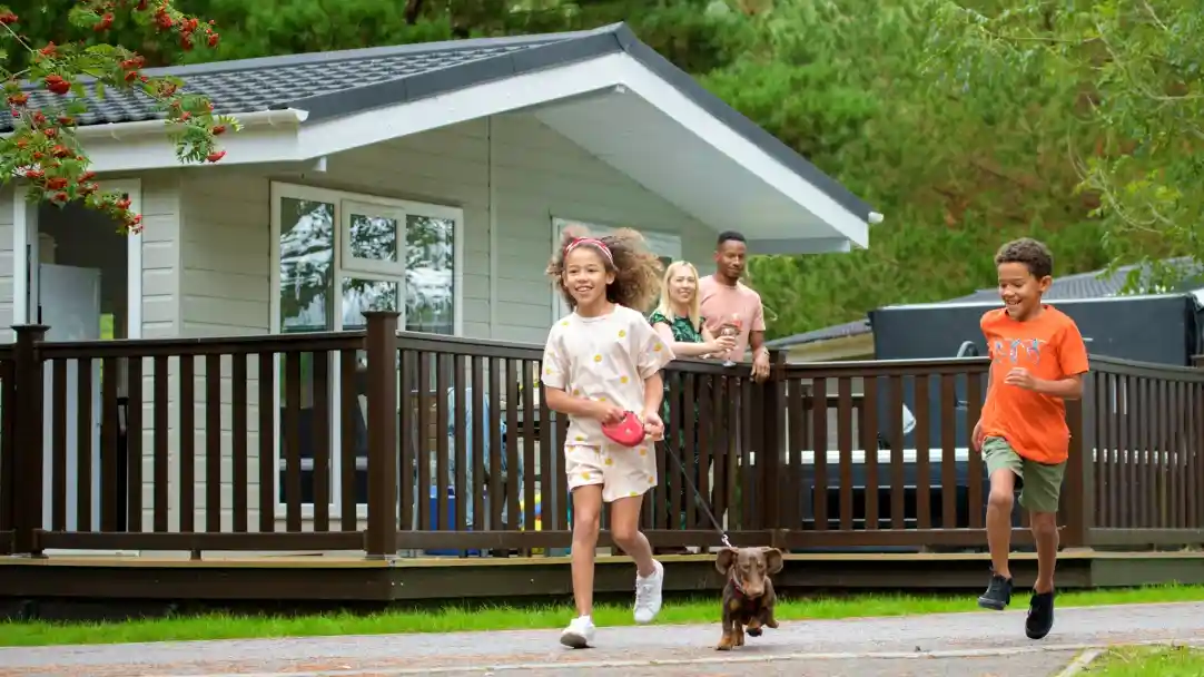 A group of children and adults enjoying a joyful moment outside a cabin, with two children running ahead while walking a dog. Lush greenery surrounds the scene, creating a cheerful atmosphere.
