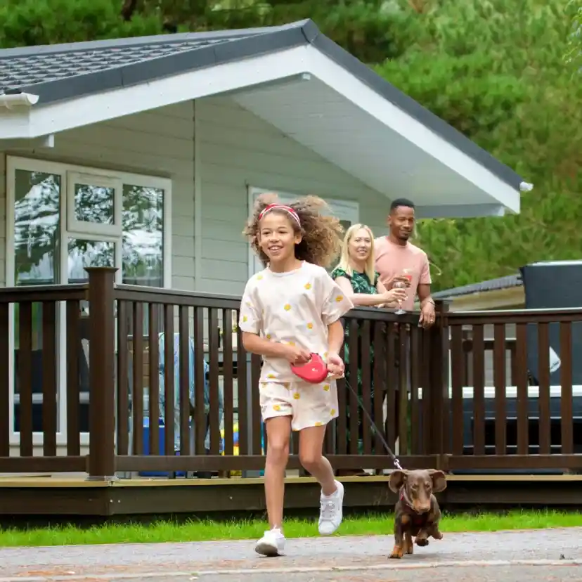A group of children and adults enjoying a joyful moment outside a cabin, with two children running ahead while walking a dog. Lush greenery surrounds the scene, creating a cheerful atmosphere.