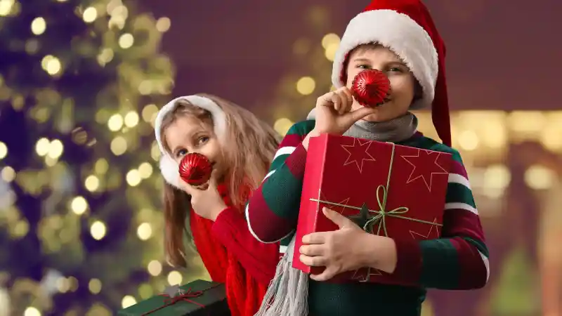 Two children in festive attire, one wearing a Santa hat, hold Christmas ornaments and gifts. Brightly decorated tree lights twinkle in the background, creating a cheerful festive holiday atmosphere.