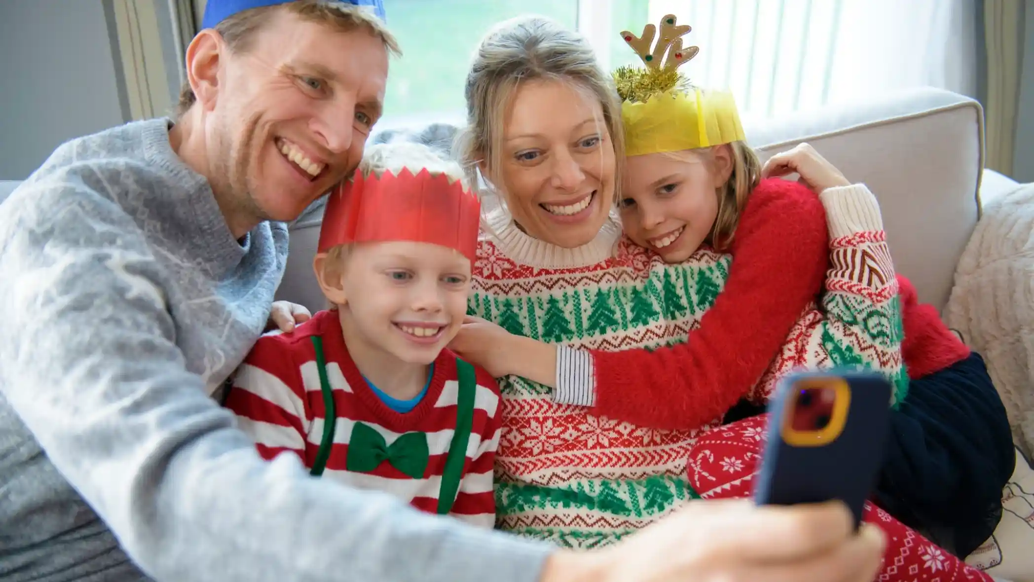 A family of four happily taking a selfie on a couch. They are wearing festive sweaters and colourful paper crowns, with two children smiling beside their parents. The setting appears cosy and cheerful, decorated for the holiday season.
