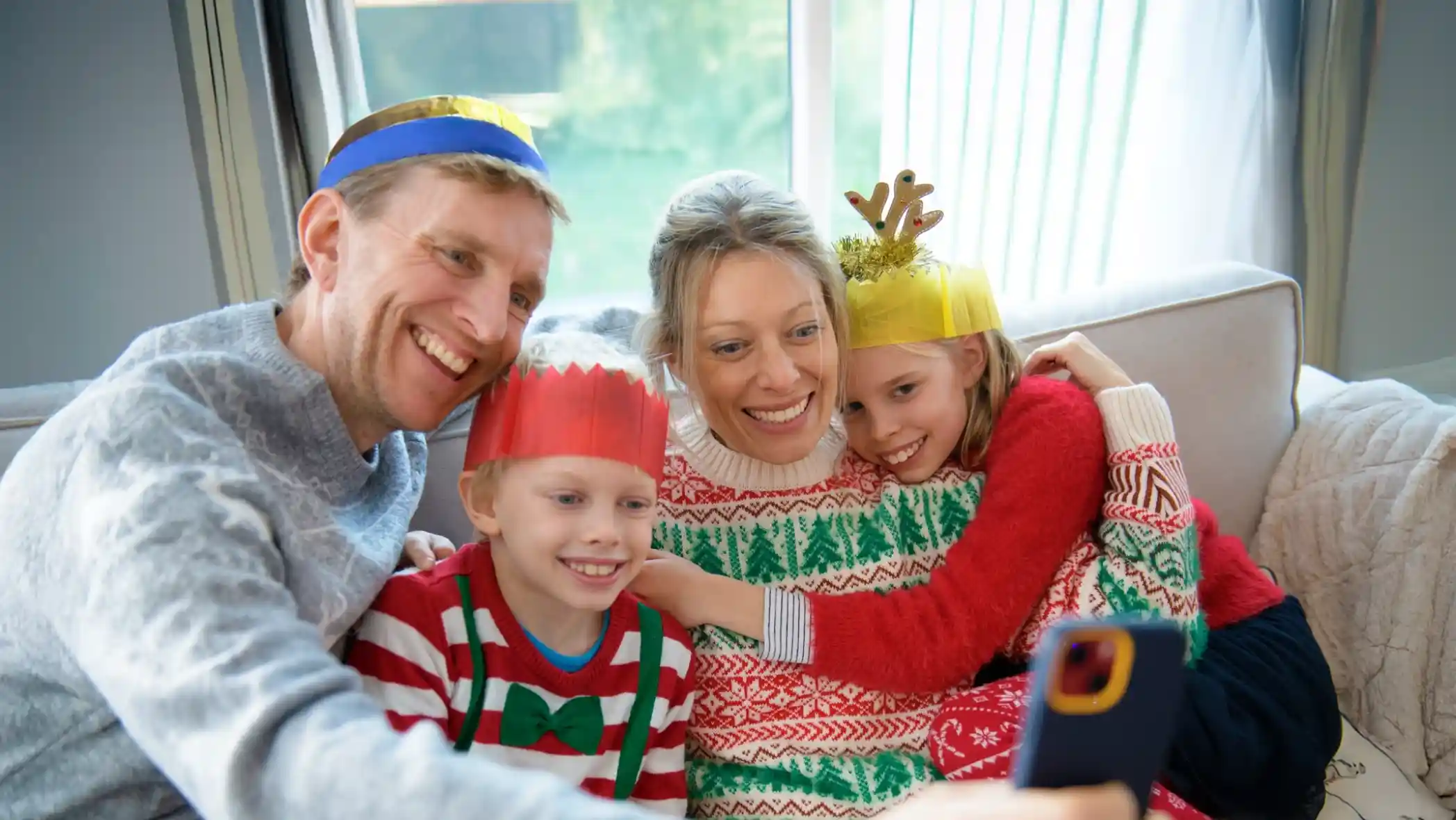A family wearing festive hats smiles together while taking a selfie. The adults and children are dressed in colourful holiday-themed sweaters, sitting on a couch with natural light streaming in.