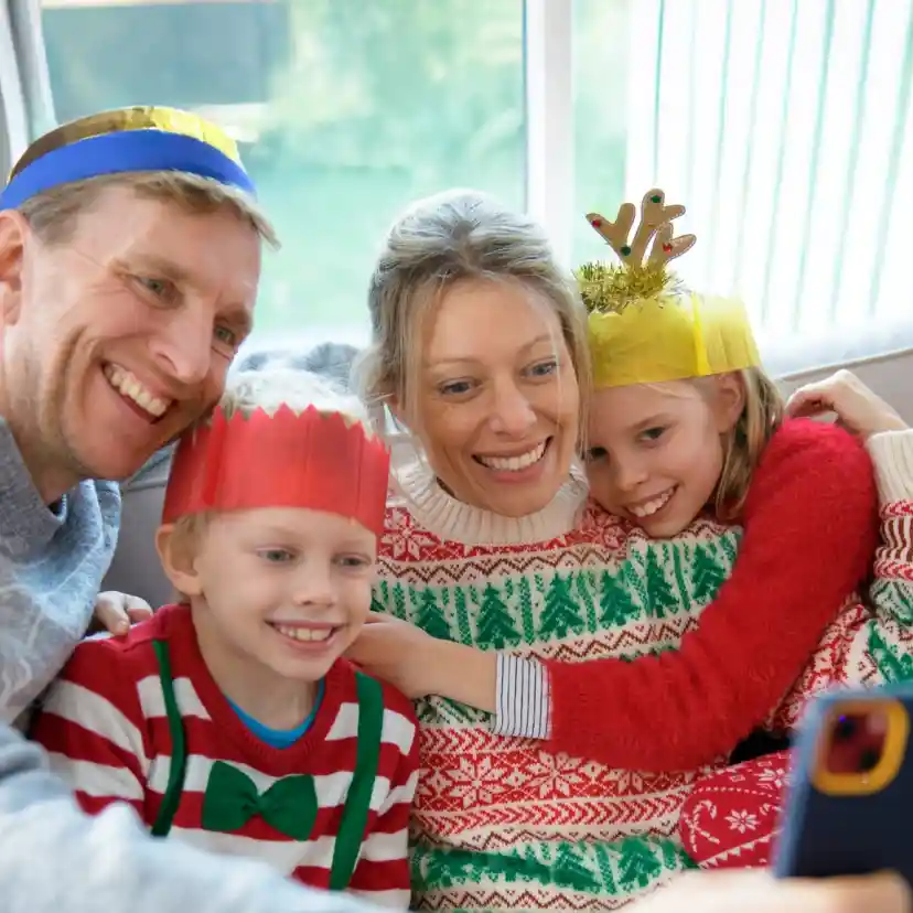 A family wearing festive hats smiles together while taking a selfie. The adults and children are dressed in colourful holiday-themed sweaters, sitting on a couch with natural light streaming in.
