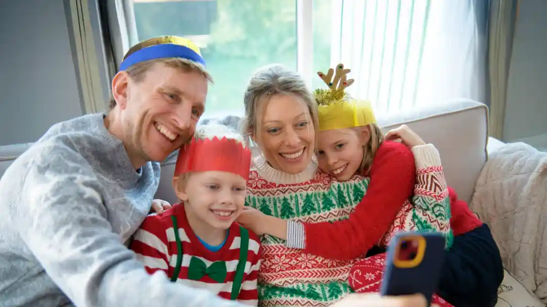 A family wearing festive hats smiles together while taking a selfie. The adults and children are dressed in colourful holiday-themed sweaters, sitting on a couch with natural light streaming in.