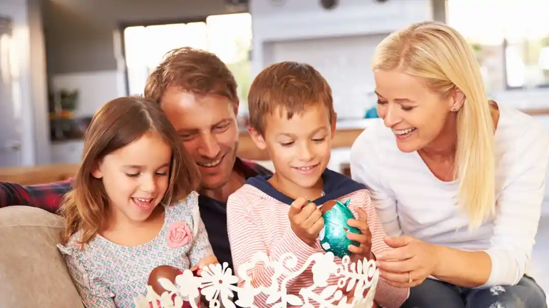 A family enjoys decorating Easter eggs together, with two children painting colorful eggs while the adults smile and engage with them. The bright, inviting home creates a warm atmosphere.