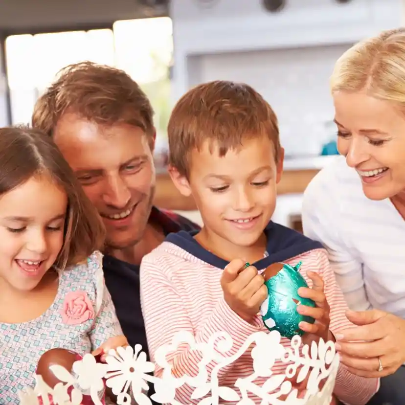 A family enjoys decorating Easter eggs together, with two children painting colorful eggs while the adults smile and engage with them. The bright, inviting home creates a warm atmosphere.
