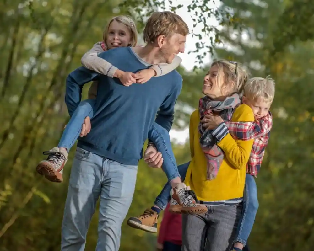 A cheerful family walks along a wooded path, with two children riding on their parents' backs. The parents smile at each other, showcasing a warm and playful atmosphere. The scene is surrounded by lush green trees, hinting at a beautiful day outdoors.