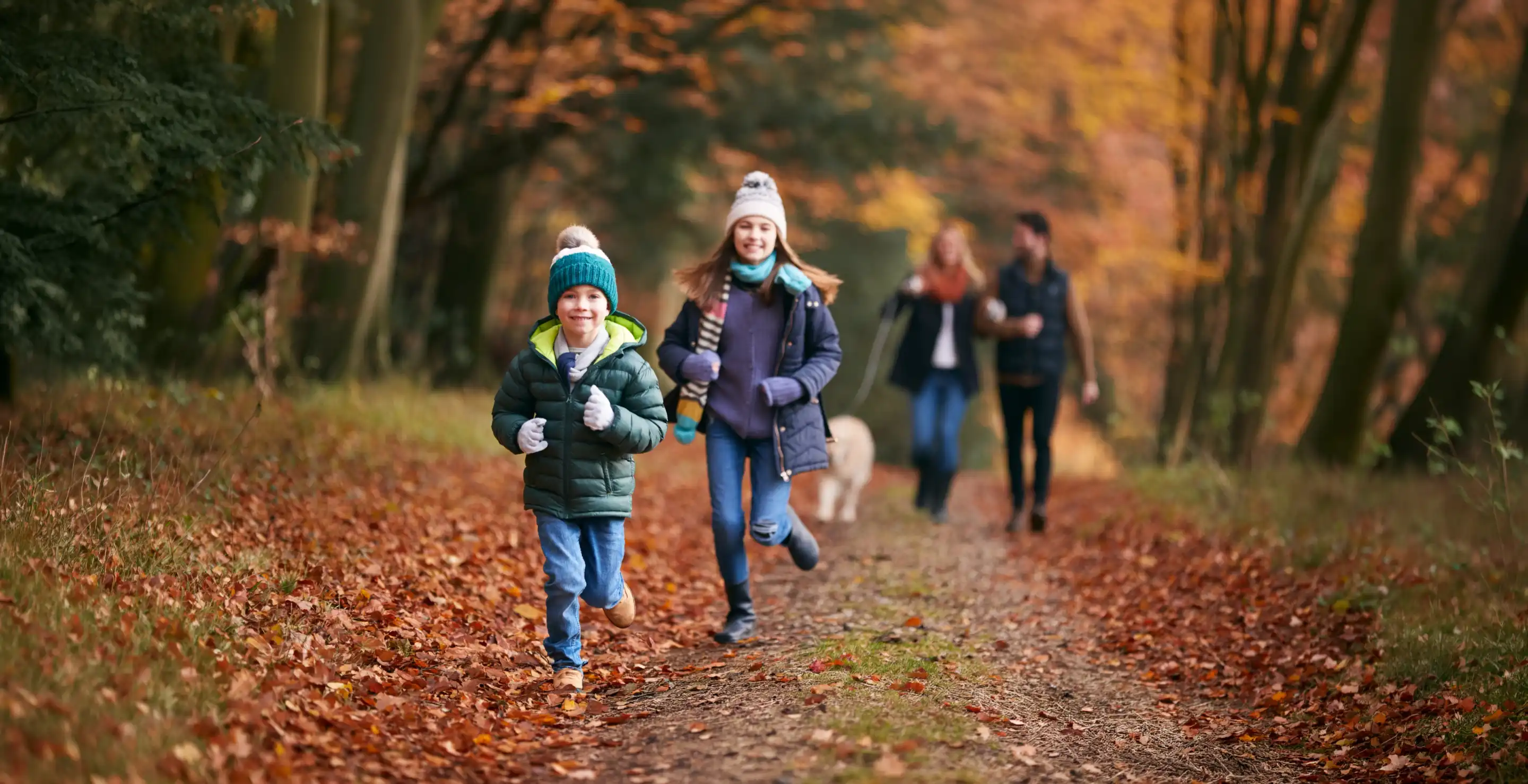 A child in a green jacket runs along a forest path covered in autumn leaves, smiling. A girl beside him wears a blue jacket and a hat, also running. In the background, two adults and a dog stroll together through the trees with colorful autumn foliage.