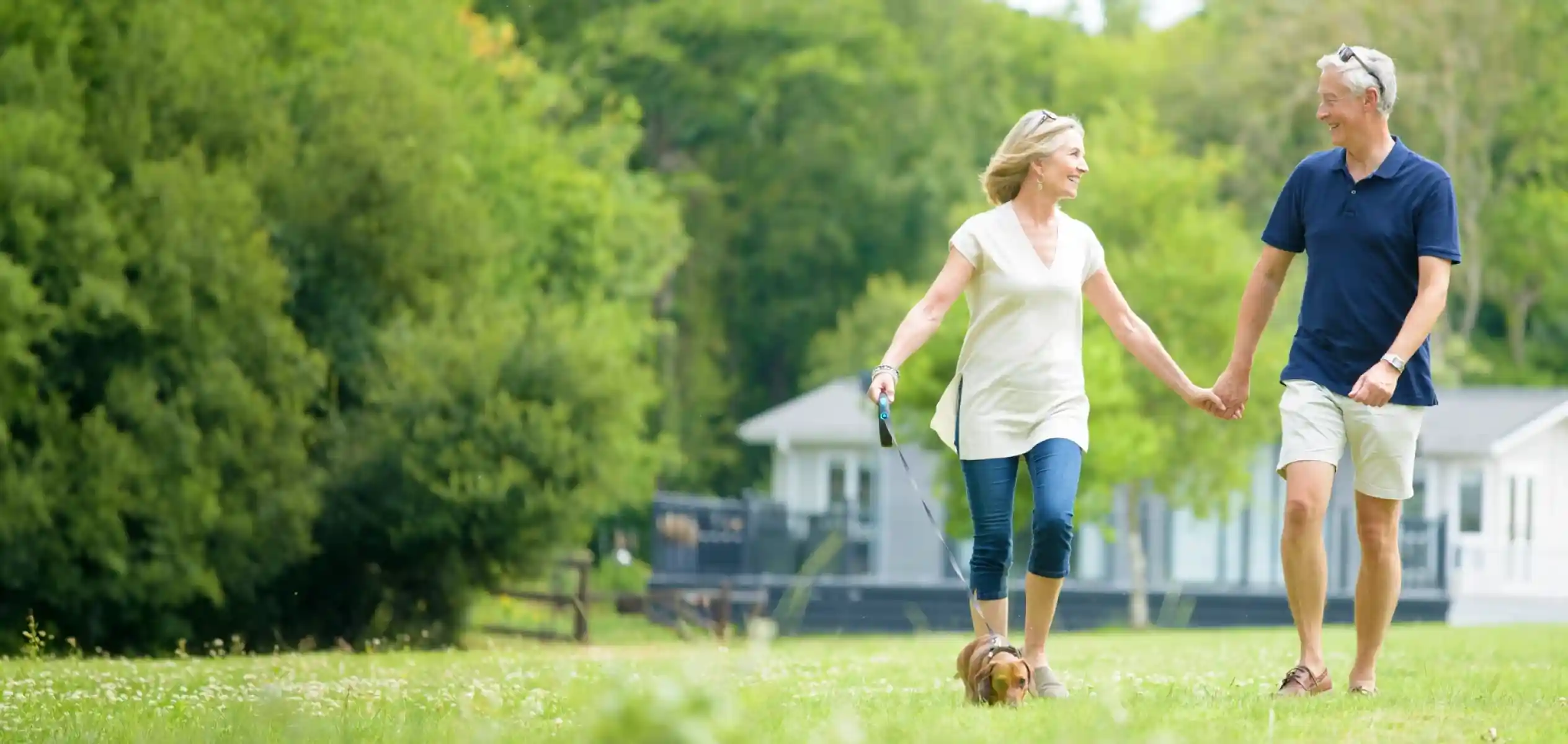 A cheerful older couple walks hand in hand through a grassy park with a small dog on a leash. Lush trees and a white holiday home are in the background.