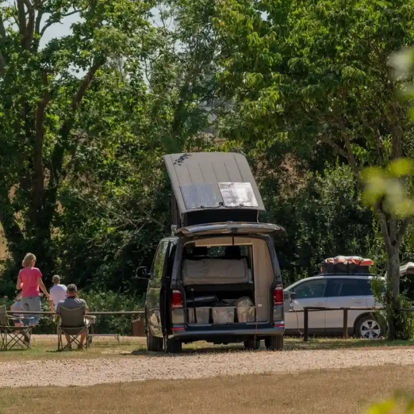 A camping scene featuring a van with a pop-up roof parked in a grassy area, surrounded by trees. Two people are sitting in chairs, enjoying the outdoors, while another person stands nearby. A second vehicle is parked in the background, also equipped for camping.