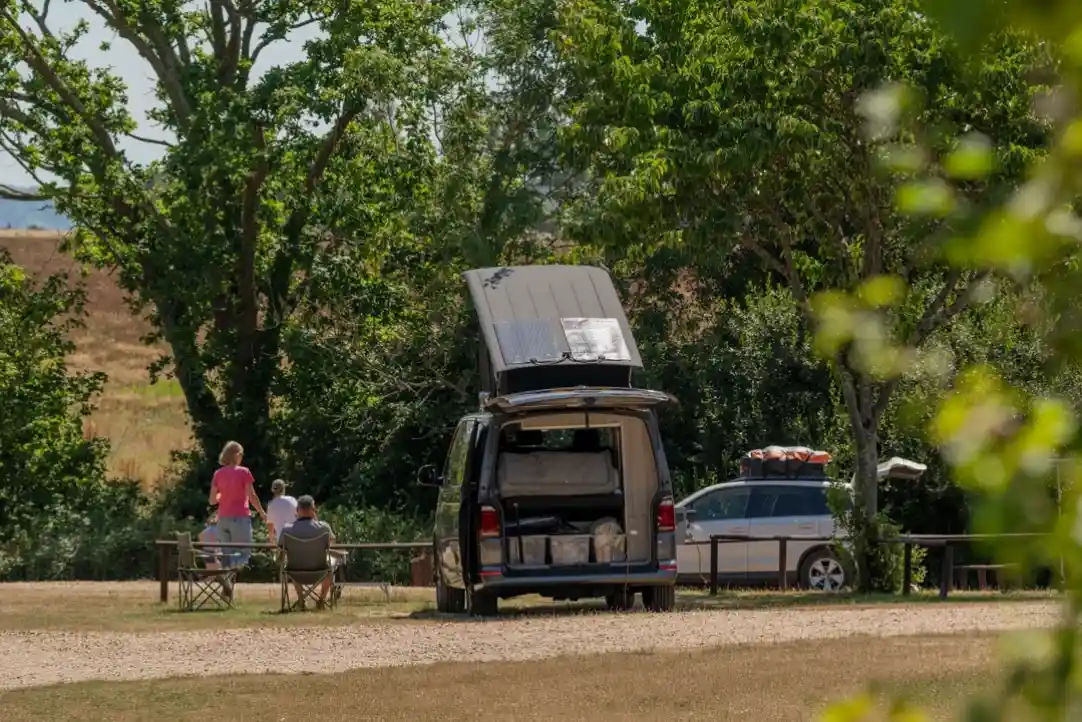 A camping scene featuring a van with a pop-up roof parked in a grassy area, surrounded by trees. Two people are sitting in chairs, enjoying the outdoors, while another person stands nearby. A second vehicle is parked in the background, also equipped for camping.