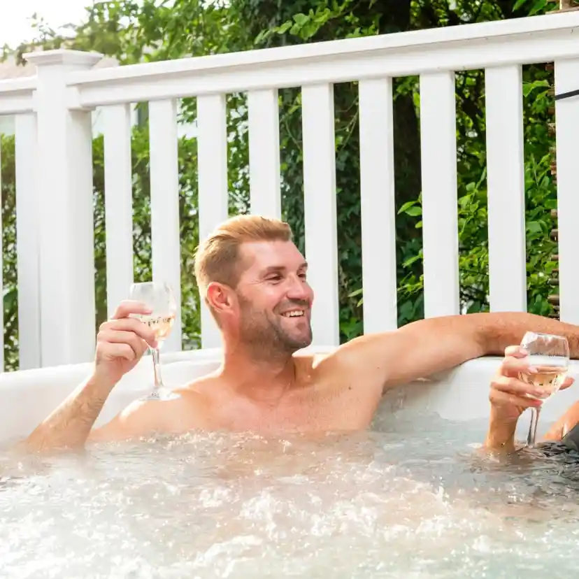 A man and a woman relax in a hot tub, holding glasses of sparkling beverages and smiling at each other, surrounded by greenery. Bright sunlight filters through the trees, creating a warm atmosphere.
