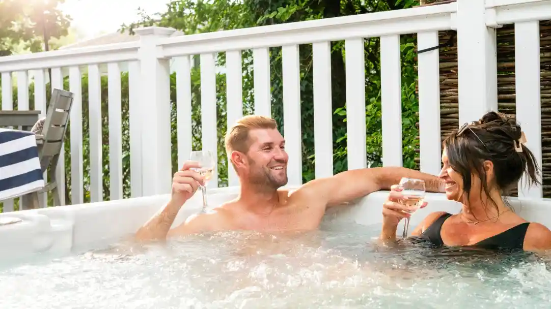 A man and a woman relax in a hot tub, holding glasses of sparkling beverages and smiling at each other, surrounded by greenery. Bright sunlight filters through the trees, creating a warm atmosphere.