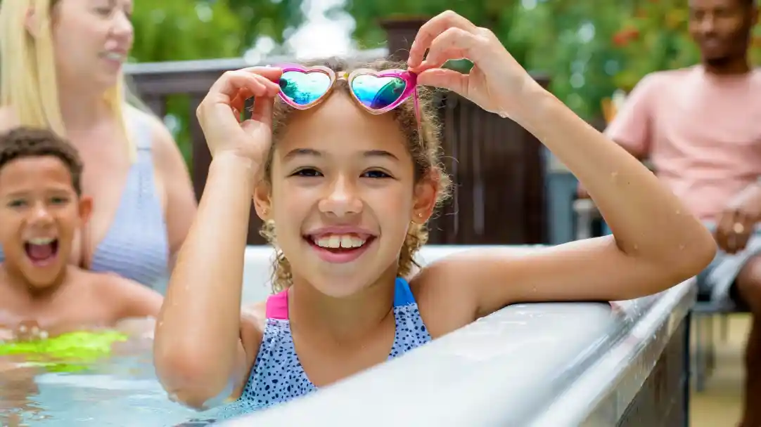 A girl with curly hair smiles brightly while holding heart-shaped sunglasses above her head in a pool. Two other children play in the background, and adults are visible nearby. The setting is sunny and cheerful, surrounded by greenery.