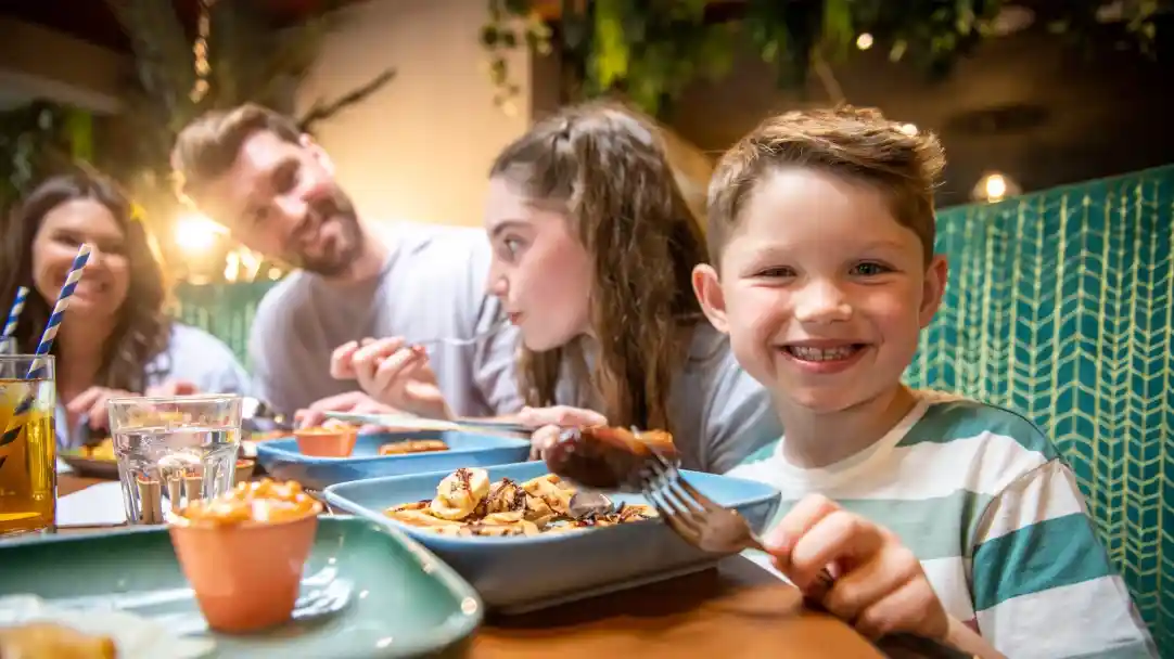 A group of people enjoying a meal at a restaurant, with a smiling young boy in the foreground holding a fork. Others are engaged in conversation while eating off various plates. The ambiance features greenery and warm lighting.
