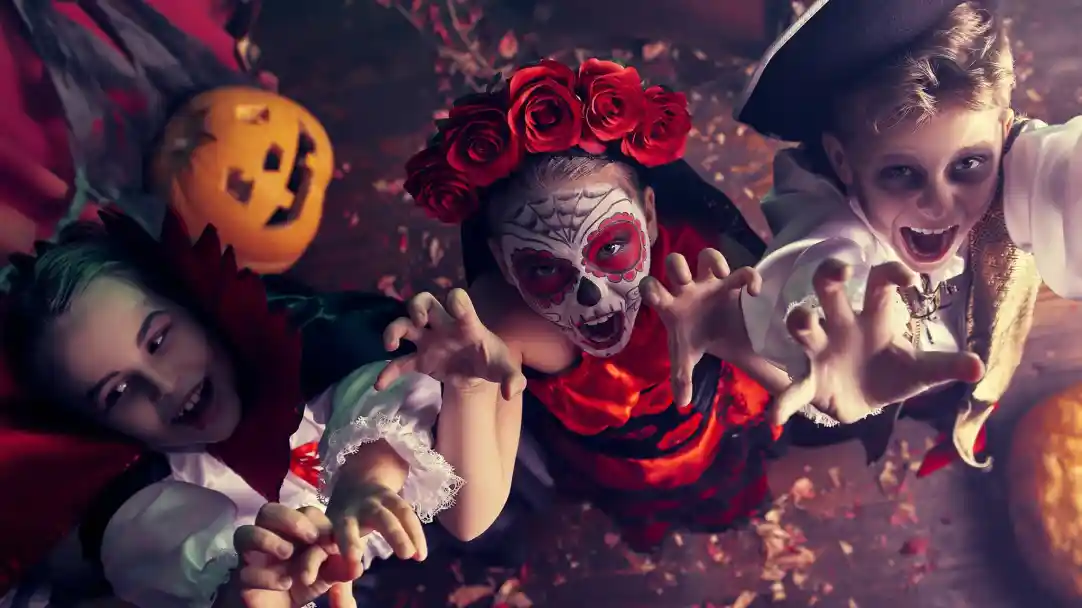 Three children dressed in Halloween costumes reach out playfully. One is a vampire with a cape, another is adorned with a face painted like a sugar skull and red roses, and the third wears a pirate outfit. A carved pumpkin is visible in the background, adding to the festive atmosphere.