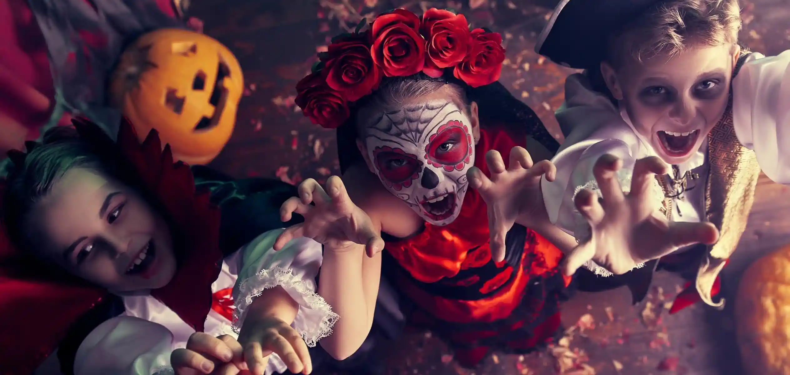 Three children dressed in Halloween costumes reach out playfully. One is a vampire with a cape, another is adorned with a face painted like a sugar skull and red roses, and the third wears a pirate outfit. A carved pumpkin is visible in the background, adding to the festive atmosphere.