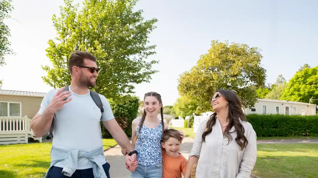 A family of four is walking together in a sunny outdoor setting. The man, wearing sunglasses and a casual outfit, laughs as he gestures while talking. A teenage girl and a young boy hold hands with him. The mother, wearing a light blouse, smiles joyfully at the man. Green trees and mobile homes are visible in the background.